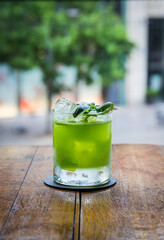 A bright green cocktail sits on a wooden bar, garnished with basil leaves and ice cubes. The background is blurred with soft, colorful bar lights.