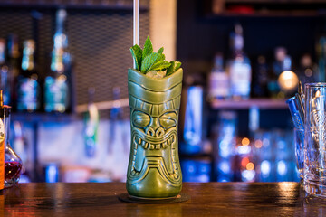 A tall, green ceramic mug with a tribal face design stands on a bar, filled with a refreshing cocktail. Fresh mint leaves garnish the drink, adding a vibrant touch.