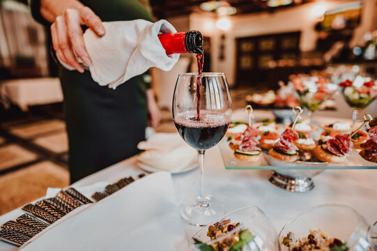 A person elegantly pours red wine into a glass at a sophisticated event. Appetizers are visible in the background, creating a refined and inviting atmosphere