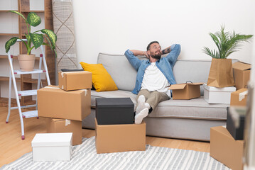 Handsome man sits comfortably on large sofa in cozy apartment thinking about completed stage of moving to house equipped with modern technologies