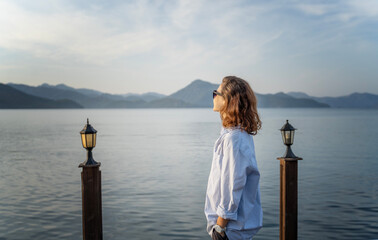 Young woman standing on the pier in the morning admiring the sea and mountain landscape