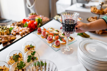 A hand holding a glass of red wine at a buffet table with various appetizers, highlighting an elegant social event. Perfect for themes of celebration, dining, and social gatherings.