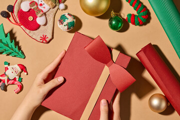 Fototapeta premium Top-down close-up photo Christmas theme showing a hand holding a red gift box. On a brown surface are Christmas themed decorations such as candy cane, Christmas balls, paper pines, and rolls of paper.