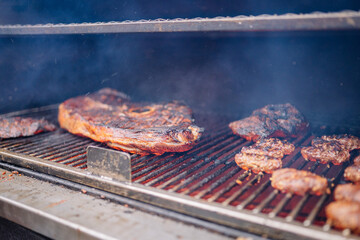 Close-up of various grilled meats cooking on a smoky barbecue grill, showing rich char marks and textures under heat..