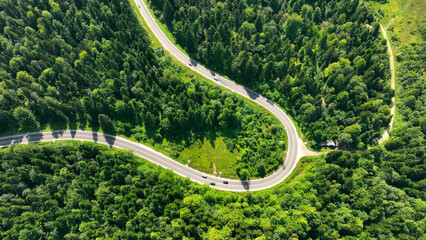 Winding mountain road through dense green forest. Aerial view of a winding road cutting through a lush, dense forest in a mountainous area, with cars navigating the curves.