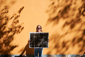 Woman holding solar panel for charging, stands against vibrant orange wall. Smiling girl wears...