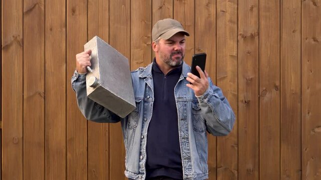 A man with an iron canister. Swearing. Telephone conversation. Bad mood. Anger. Wooden house in the background. Dressed warmly. Bearded. Denim jacket and baseball cap. Rural area.