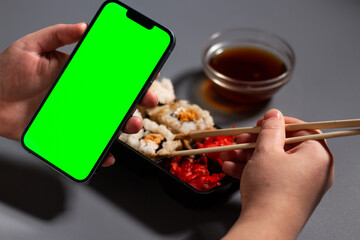 Female using smartphone while eating sushi rolls at home. Smartphone with green screen with space for text. Smartphone in hands with served table in background.