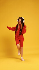 Full-length shot of girl in red sweatshirt with headphones, playfully dancing against yellow studio background. Concept of childhood, lifestyle, expressions, emotions, trendy. Ad