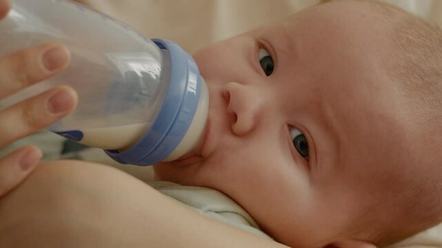 Shot of infant drinking milk while in mother's hands. Newborn baby boy eats milk from bottle. Family and child care.
