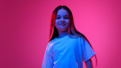 Fototapeta premium Young fashionista. Little girl, dressed white t-shirt smiling and looking at camera in neon light against vivid gradient background. Concept of childhood, lifestyle, expressions, trendy. Ad