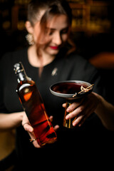 Focus on glass of drink and bottle of whiskey in hands of young pretty female bartender