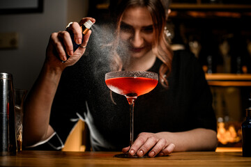 Smiling female bartender spraying brown cocktail in tall stemmed glass
