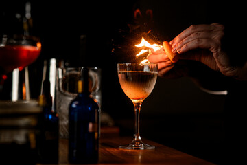 Focus on goblet with clear liquid over which female hands of bartender squeeze a piece of zest