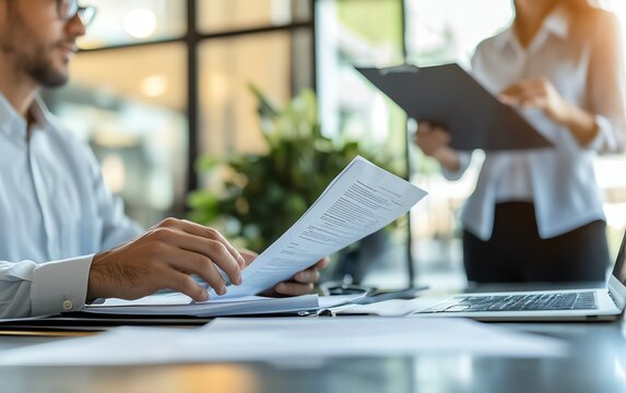 An office interview setup with one person sitting at a desk with a laptop and papers, while the other stands with documents, in a clean and modern office space - Powered by Adobe