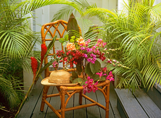 Hibiscus, orchids and bougainvillea still life in a vase on a cane chair with sun hat surrounded by green palm leaves in a conservatory