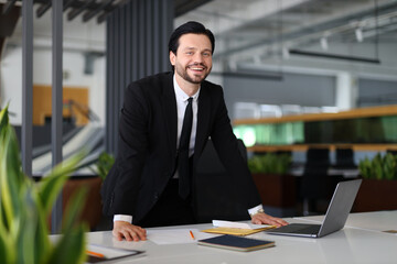 A man in a suit and tie stands in front of a desk with a laptop and a stack of papers. He is smiling and he is in a professional setting