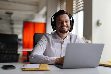 A man wearing headphones is sitting at a desk with a laptop. He is smiling and he is enjoying his work