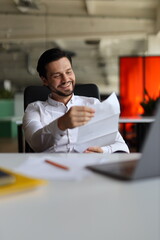 A man is sitting at a desk with a piece of paper in his hand. He is smiling and he is happy