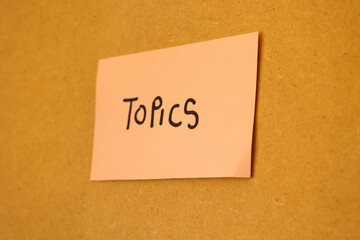 Close up of pink note stuck on a board with written message saying topics, concept of notes and topics