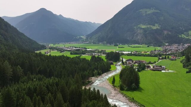Aerial dolly descends along quaint town nesteled in valley leading to Reinbach waterfalls cascading down through lush forested landscape in Italy