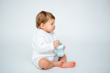 A baby boy with a bottle of milk on a white isolated background with a place for text is sitting in white knitted clothes. A small happy child drinks milk, space for text