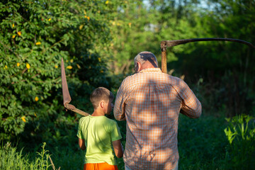 A man and a boy, father and son, work together in the field. The father with a scythe in his hands stands with his back to the camera.