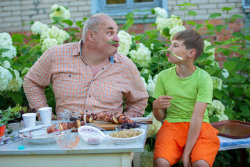 A grandfather and his grandson are happily eating shashlik, chatting and playing around in the yard of a village house. A man and a boy made moustaches using green onions and are laughing.