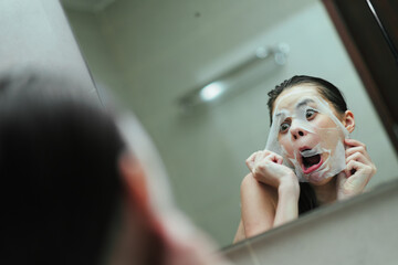 Humorous Asian woman making funny faces while applying a facial mask in the bathroom mirror. Self-care and skincare routine concept.