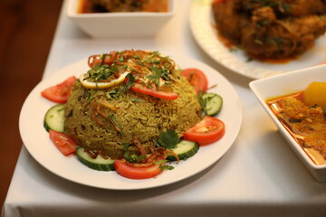 Fragrant green rice garnished with fresh herbs, tomato, and cucumber sits on a white tablecloth, accompanied by flavorful side dishes.