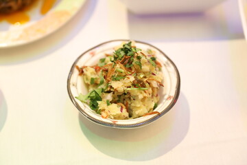 Dish of creamy potato salad, garnished with fresh cilantro and sliced almonds, served in a traditional bowl. The background features aromatic dishes.