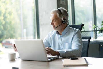 Fototapeta premium Smiling businessman using headset when talking to customer. Tech support manager in headset consulting a client