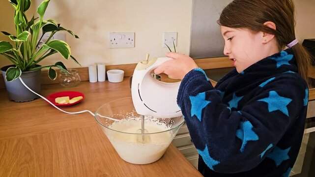 Child in Blue Star-Patterned Robe Uses Mixer to Beat Eggs for Cream Preparation in Cozy Kitchen During Day. Mixer whips sugar and eggs in glass bowl in kitchen. Cooking and baking concept