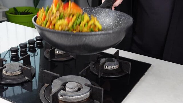 Close up of Chef cook hands cooking and toss roasted pasta, onion, pepper, carrot, vegetables, mushrooms, green peas, beans, garlic in frying wok pan on gas stove. Flying food levitation.