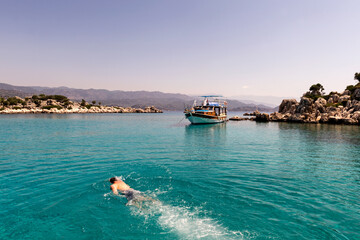 Obraz premium A serene image of a person swimming in the crystal-clear turquoise waters near a boat in Kaş, Turkey, surrounded by rocky islets and distant mountains