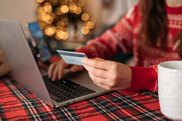 Woman in cozy sweater holding credit card and shopping online on a laptop, holiday lights and plaid...