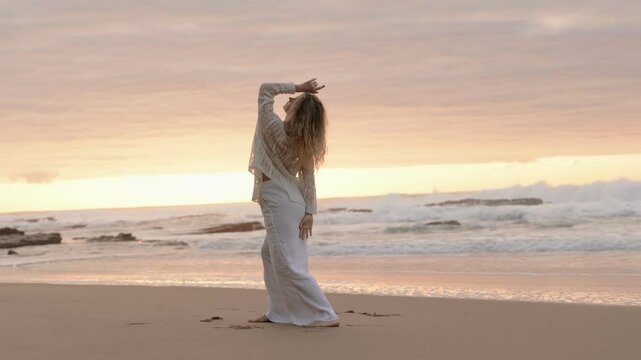 An elegant dancer demonstrates a contemporary dance move on a tranquil beach at sunset, dressed in white.