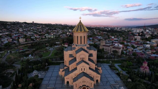 Aerial Drone Footage Showcases The Stunning Holy Trinity Church In Tbilisi, Georgia During A Vibrant Summer Evening. The Camera Circles The Church, Highlighting Its Majestic Dome Against A Bright Sky.