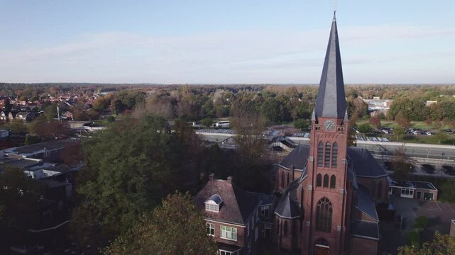 Parish St. Antonius of Padua church in Dutch town Nijverdal seen from above on a sunny day with the tower rising above countryside landscape revealing train station and tunnel in the background
