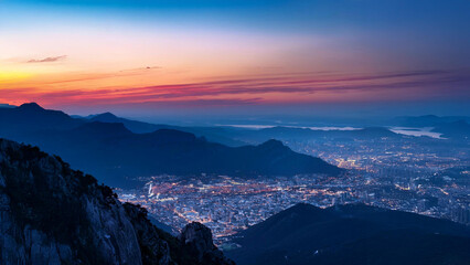 Mountain top at twilight, with a glowing city far below, surrounded by mountains and bathed in soft blue and pink hues