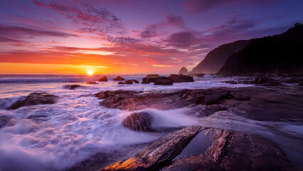 Obraz premium Sunrise on a rocky coastline with waves crashing, orange and purple skies reflected in tide pools, and distant cliffs