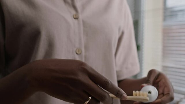 Cropped shot of unrecognizable Black woman taking natural bamboo toothbrush from stylish holder on bathroom countertop and squeezing toothpaste on it while doing morning routine at home