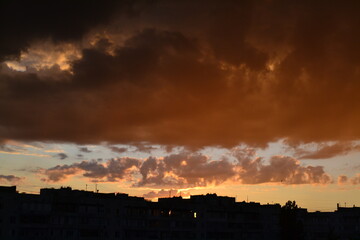 Bright unusual sunset city landscape orange pink rays above cityscape. Brutalist panel buildings appear as black silhouettes against a fiery orange-yellow sky dramatic lighting dark rain storm clouds