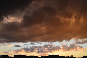 Bright unusual sunset city landscape orange pink rays above cityscape. Brutalist panel buildings appear as black silhouettes against a fiery orange-yellow sky dramatic lighting dark rain storm clouds