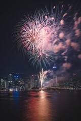 Colorful fireworks display over a city skyline at night, with the city's buildings and water reflections visible