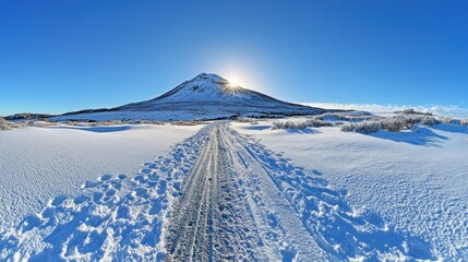 A snowy road leads towards a snow-capped mountain with the sun shining brightly in the sky.