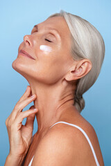 Side profile portrait of a serene mature 50s woman with gray hair, applying skin care moisturizer...
