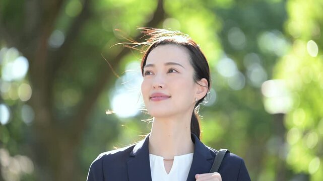 逆光の美しい新緑や緑の背景でビジネススーツを着た笑顔の美人な営業マン女性　見上げるクローズアップのコピースペース　未来や転職のイメージのスロー動画