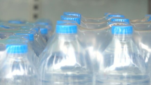 A row of plastic bottles of water are lined up on a shelf. The bottles are clear and blue
