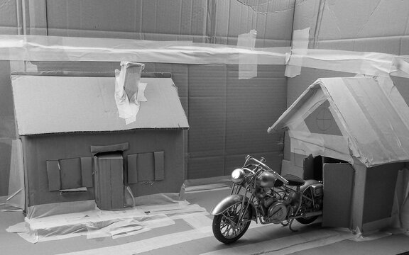Historisch, Oldtimer, Motorrad Model, &auml;hnlich einer Harley Davidson, f&auml;hrt aus einer Garage in Karton Modell-Landschaft in Schwarz Weiss
